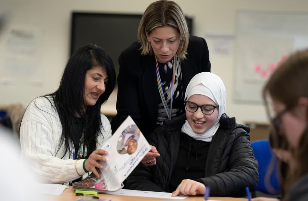 Two adults and a young person smiling as they look through a booklet together at a classroom table.