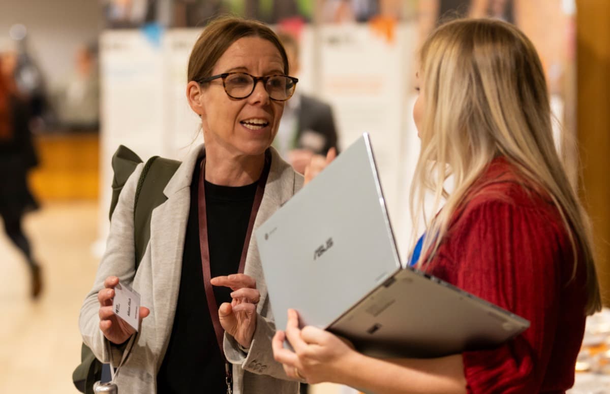 Two people in conversation at an event, one holding a laptop.